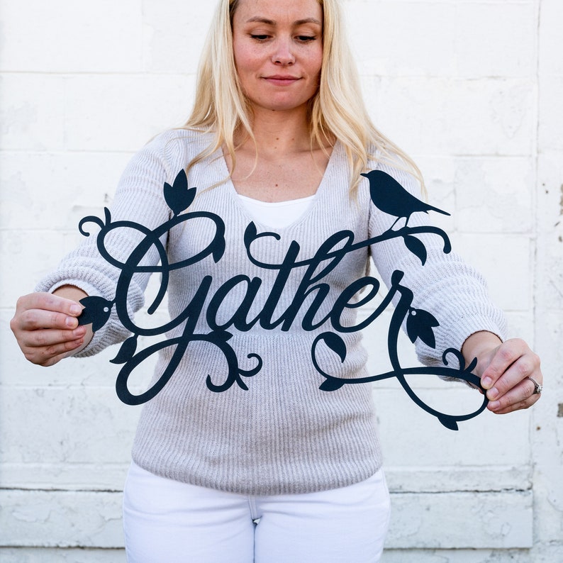 May include: A woman holds a black metal sign with the word "Gather" in a decorative script font. The sign features a bird perched on a branch with leaves.
