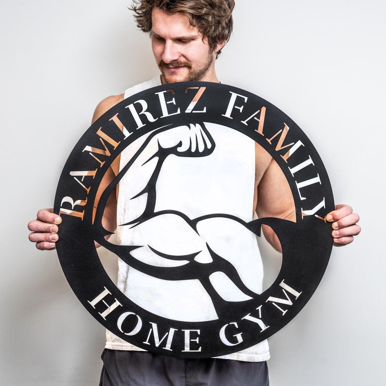 K&ouml;nnte beinhalten: Schwarzes Metallschild mit einer wei&szlig;en Silhouette eines sich anstrengenden Bizepses. Das Schild tr&auml;gt die Aufschrift "Ramirez Family Home Gym" in kupferfarbener Schrift.