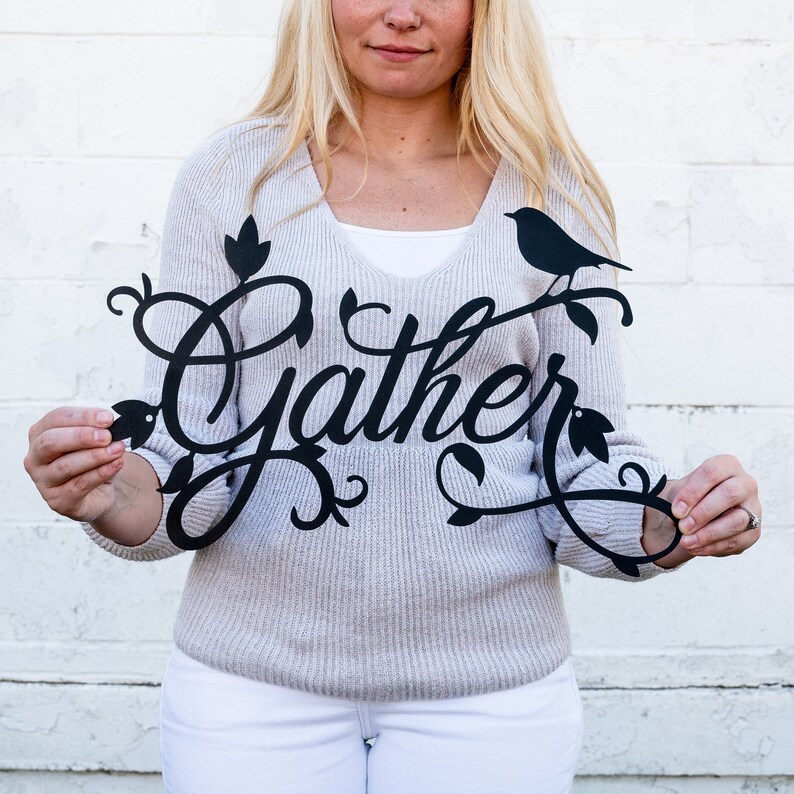 May include: A black metal wall decor featuring the word "Gather" in a decorative script font. The word is surrounded by stylised leaves and a small bird silhouette.