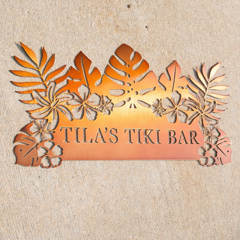 Puede incluir: Un letrero de bar tiki de metal con un dise&ntilde;o de hojas tropicales y el texto "Tila's Tiki Bar".