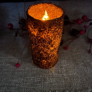 May include: A brown and gold textured candle with a flickering flame inside. The candle is sitting on a beige textured surface.