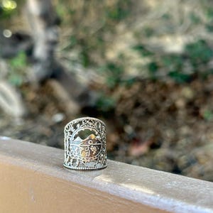May include: Silver-toned ring with a mountain and foliage design. The ring has an openwork design with intricate details. The ring is approximately 1 inch in height and is set against a blurred natural background.