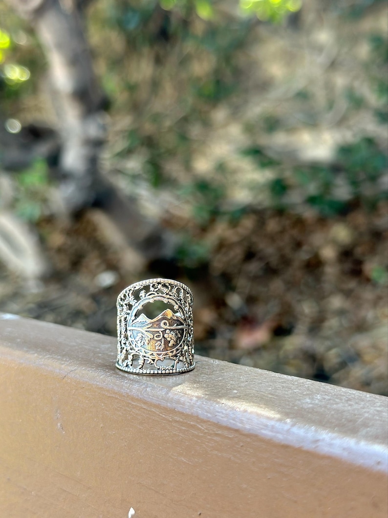 May include: Silver-toned ring with a mountain and foliage design. The ring has an openwork design with intricate details. The ring is approximately 1 inch in height and is set against a blurred natural background.