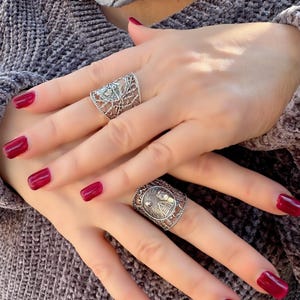 May include: Two silver rings with intricate designs are displayed on a person's hands. The rings feature detailed patterns and are worn on different fingers. The person's nails are painted a deep red color, complementing the silver rings.