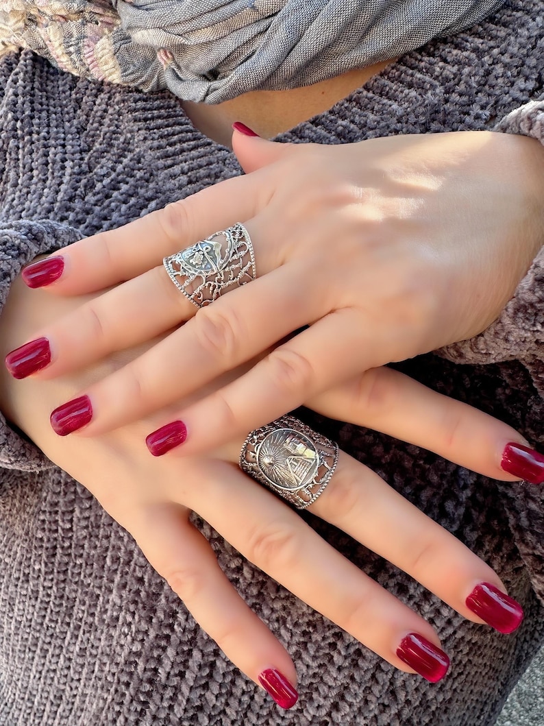 May include: Two silver rings with intricate designs are displayed on a person's hands. The rings feature detailed patterns and are worn on different fingers. The person's nails are painted a deep red color, complementing the silver rings.