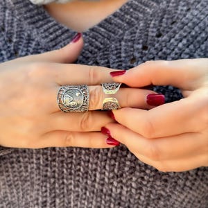 May include: Close-up of three silver rings with intricate designs. The rings are worn on the fingers. The nails are painted a deep red. The rings have detailed patterns, including a rectangular ring with a detailed design.