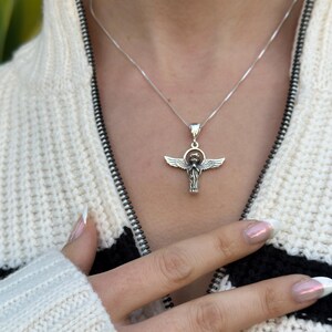 May include: A silver necklace with an angel pendant. The angel is depicted with wings outstretched and a halo. The necklace is worn by a person with a white sweater.
