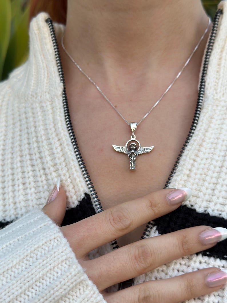 May include: A silver necklace with an angel pendant. The angel is depicted with wings outstretched and a halo. The necklace is worn by a person with a white sweater.