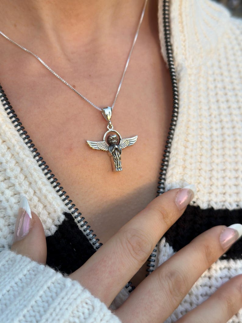 May include: A silver chain necklace with a pendant featuring a detailed angel with outstretched wings. The angel is holding a sphere in its hands.