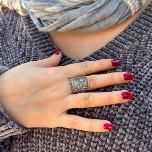 May include: A silver-toned ring with a detailed design featuring a central emblem and intricate patterns. The ring is worn on a hand with red nail polish, set against a gray sweater and a light blue scarf.