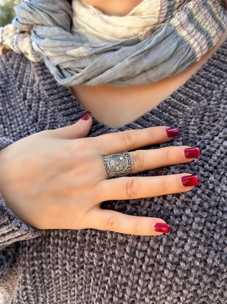 May include: A silver-toned ring with a detailed design featuring a central emblem and intricate patterns. The ring is worn on a hand with red nail polish, set against a gray sweater and a light blue scarf.
