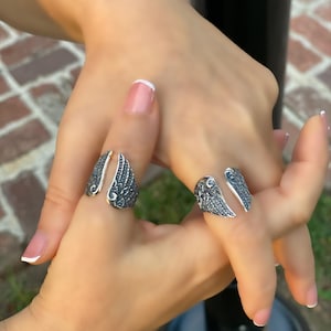 May include: Two silver rings with detailed wing designs. The rings are worn on the index and middle fingers of a hand.