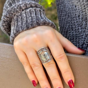 May include: A silver-colored ring with an intricate openwork design. The ring features a central motif with a mountain and bird silhouette. The ring is worn on a hand with red nail polish, and the person is wearing a gray sweater.