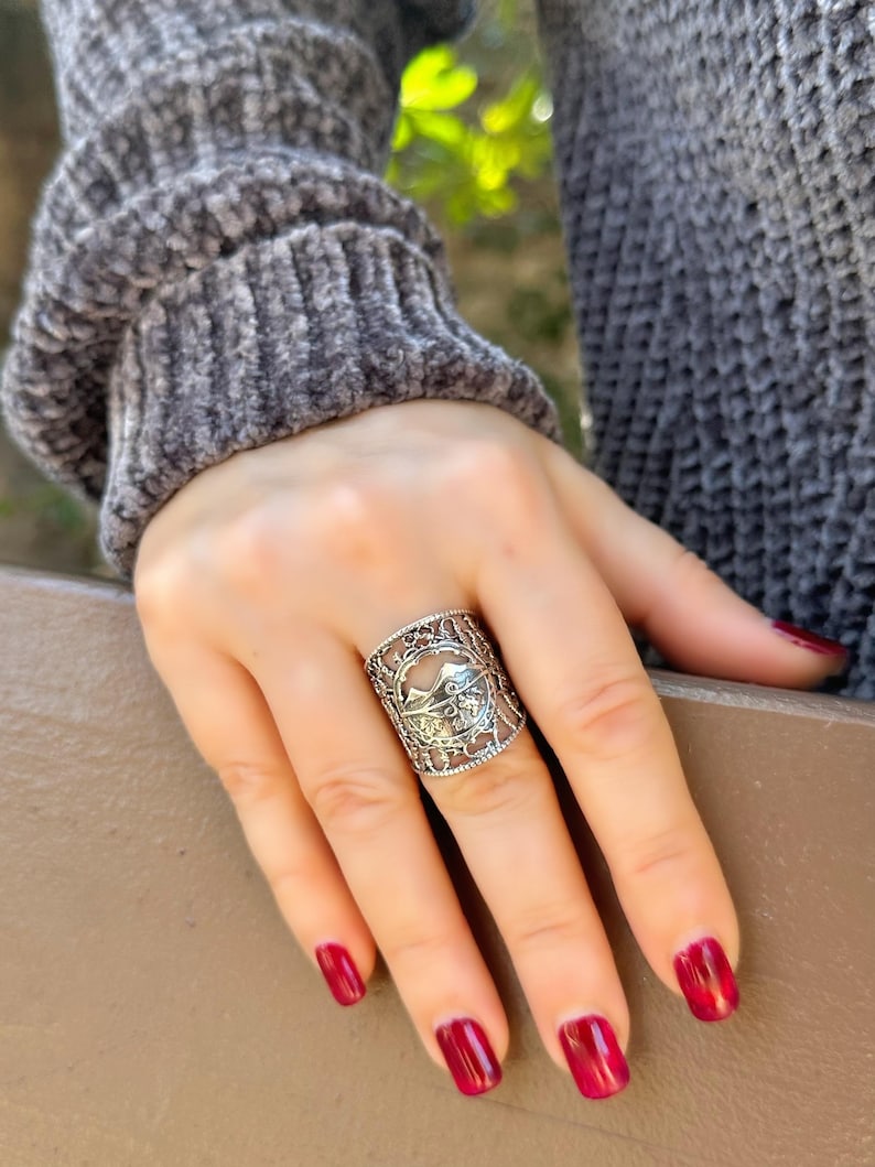 May include: A silver-colored ring with an intricate openwork design. The ring features a central motif with a mountain and bird silhouette. The ring is worn on a hand with red nail polish, and the person is wearing a gray sweater.