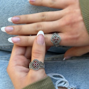 May include: Two silver rings with intricate designs and red gemstones. The rings are worn on the fingers of a person's hand.