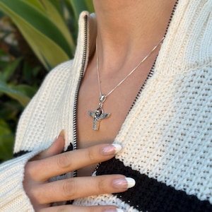 May include: A silver necklace with a pendant featuring an angel with outstretched wings. The pendant is on a silver chain and is worn by a person with a white sweater.