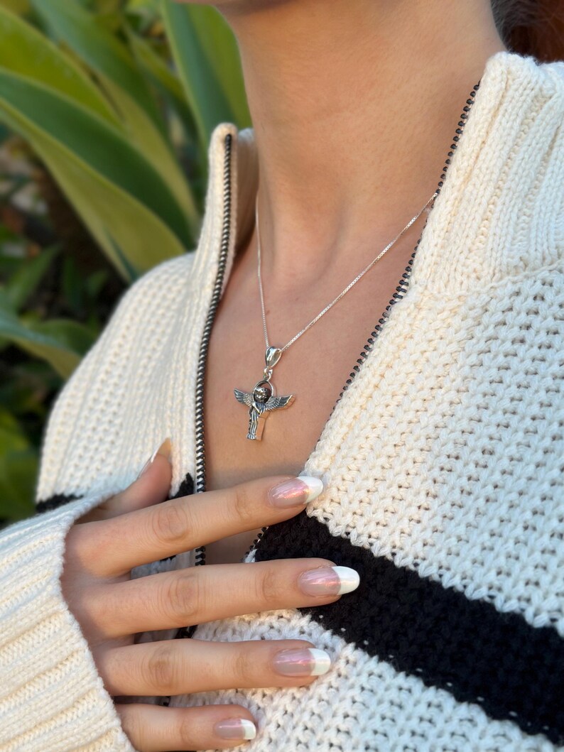 May include: A silver necklace with a pendant featuring an angel with outstretched wings. The pendant is on a silver chain and is worn by a person with a white sweater.