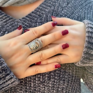 May include: A silver filigree ring with an ornate design is worn on a finger. The ring is the focal point, set against a backdrop of a gray sweater and hands with red nail polish. A gray scarf is visible in the background.