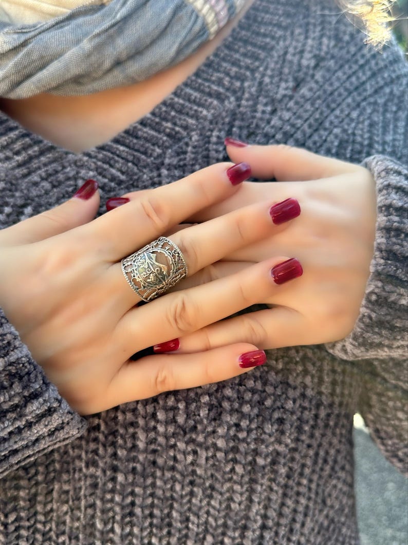 May include: A silver filigree ring with an ornate design is worn on a finger. The ring is the focal point, set against a backdrop of a gray sweater and hands with red nail polish. A gray scarf is visible in the background.