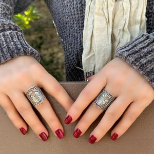 May include: Two silver rings with intricate designs are displayed. The rings are worn on fingers with red nail polish. The background includes a gray sweater and a patterned scarf.