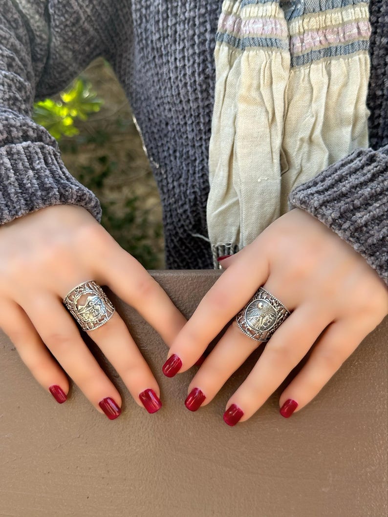 May include: Two silver rings with intricate designs are displayed. The rings are worn on fingers with red nail polish. The background includes a gray sweater and a patterned scarf.
