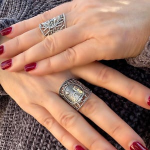 May include: Two silver rings with intricate designs are displayed. One ring features a floral pattern, while the other showcases a sun and mountain scene. The rings are worn on fingers with red nail polish, set against a gray background.