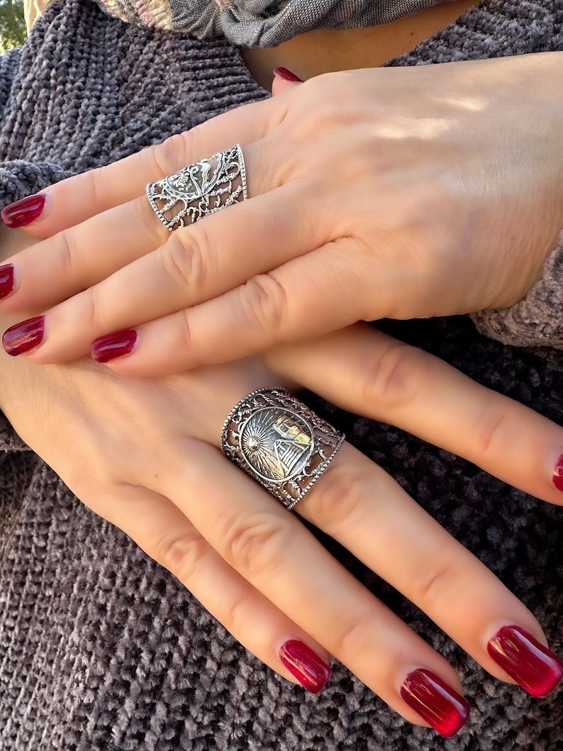 May include: Two silver rings with intricate designs are displayed. One ring features a floral pattern, while the other showcases a sun and mountain scene. The rings are worn on fingers with red nail polish, set against a gray background.