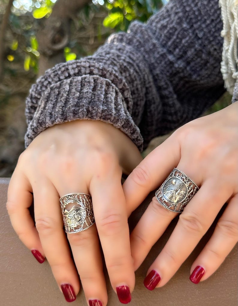 May include: Two silver rings with intricate filigree designs. Each ring features a central circular motif with a detailed image, surrounded by openwork patterns. The rings are worn on fingers with red nail polish.