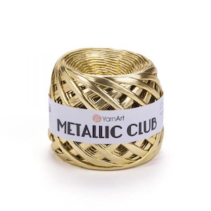 May include: A ball of metallic gold yarn wrapped in a white paper label. The label features the text YarnArt and METALLIC CLUB in black lettering. The yarn has a smooth, reflective surface and is wound into a cylindrical shape with a flat top.