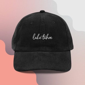 Pode incluir: Boné de beisebol de veludo cotelê preto com o texto bordado "lake tahoe" em escrita branca. O boné tem uma aba curva e um botão superior. O fundo é um gradiente de rosa e branco.