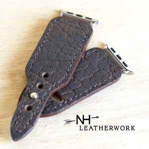 May include: Two brown leather watch bands with a textured surface and silver metal clasps. The bands are designed for Apple Watches. The bands are stacked on top of each other. The text "NH- LEATHERWORK" is visible in the bottom right corner of the image.