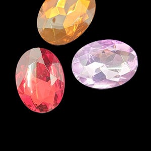 Peut inclure: Trois pierres précieuses ovales à facettes disposées sur un fond noir. Une pierre est orange vif, une est rouge foncé et la troisième est d'un mauve pâle. Chaque pierre précieuse présente une surface à facettes multiples qui reflète la lumière.