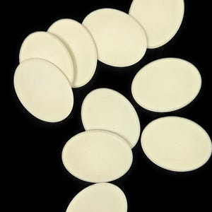 May include: A collection of ten smooth, off-white oval-shaped discs arranged in a cluster. Each disc features a uniform, matte surface texture and rounded edges, presented against a solid black background to highlight their pale, neutral colour.
