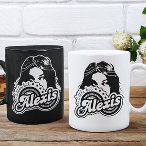 May include: Two ceramic coffee mugs with a black and white design. The design features a woman with long hair and the text "a little bit Alexis".