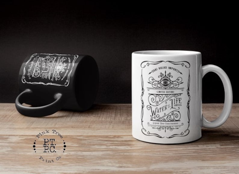 May include: Two ceramic mugs with black and white designs. The mug on the left is black with white text and a design. The mug on the right is white with black text and a design. The text on the mug on the right reads "Chai Wild Imports - Limited Edition - Water of Life - Liquid Mist Concentration".