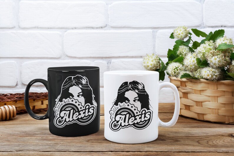 May include: Two ceramic coffee mugs with a black and white design. The design features a woman with long hair and the text "a little bit Alexis".