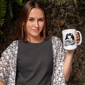 May include: A white ceramic mug with a black and white graphic design featuring a woman's face and the text "Little bit Alexis".