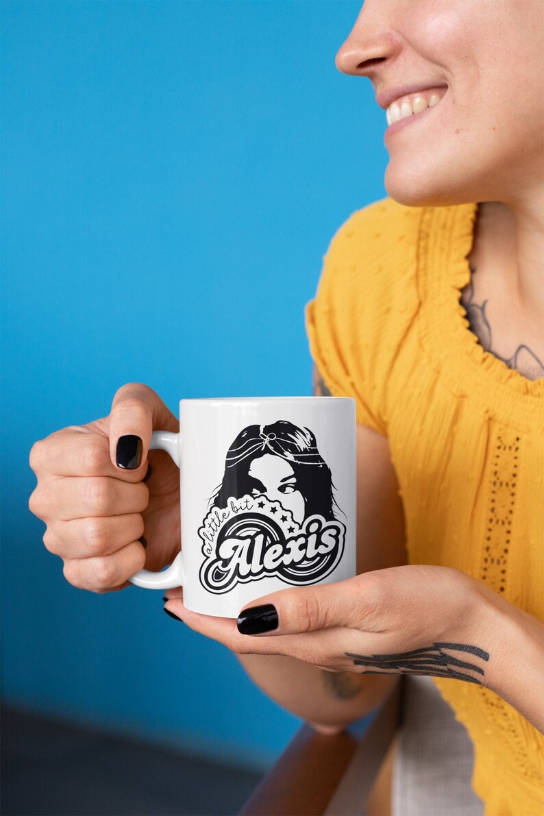 May include: White ceramic mug with a black and white design. The design features a woman's face with a headband and the text "a little bit Alexis".