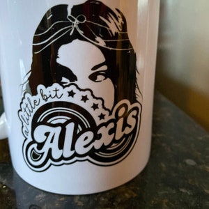 May include: White ceramic mug with a black and white design. The design features a woman's face with long hair, a circle with the word "Alexis" in a retro font, and the text "a little bit" above the circle.