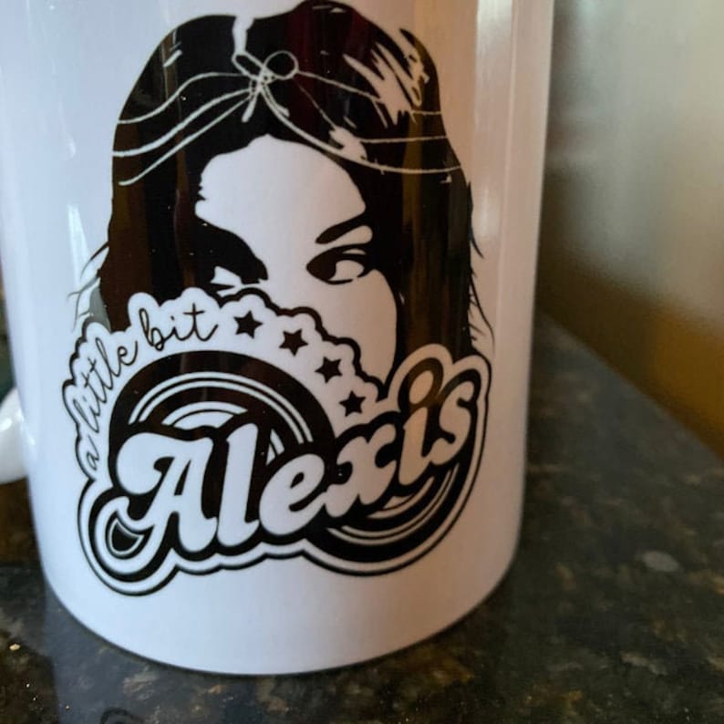 May include: White ceramic mug with a black and white design. The design features a woman's face with long hair, a circle with the word "Alexis" in a retro font, and the text "a little bit" above the circle.