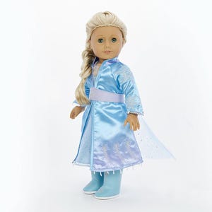 May include: A doll wearing a light blue dress with a matching cape and boots. The dress has a silver belt and snowflake embroidery. The doll has long blonde hair and blue eyes.
