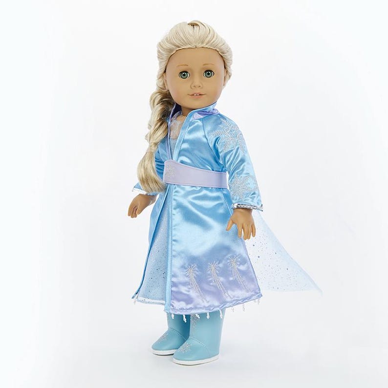 May include: A doll wearing a light blue dress with a matching cape and boots. The dress has a silver belt and snowflake embroidery. The doll has long blonde hair and blue eyes.