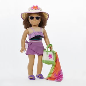 May include: A doll wearing a purple and white swimsuit, a straw hat with a flower, sunglasses, and sandals. The doll is holding a green and white beach bag and a colorful beach towel.