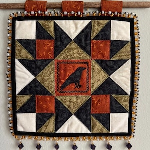 May include: A square quilted wall hanging featuring a central orange square with a black crow silhouette. The quilt uses a star pattern with black, white, and olive green fabric pieces. The border is decorated with black, white, and gold beads.