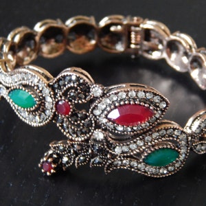 May include: A gold-toned bracelet with intricate floral designs and colourful gemstones. The bracelet features red, green, and clear stones set in a silver-toned metal.
