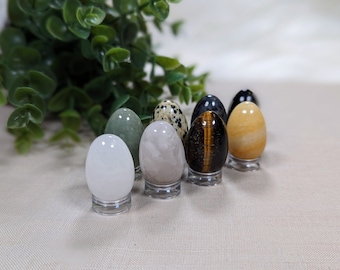 May include: Eight polished gemstone eggs on small clear stands. The eggs are in various colors and patterns, including white, green, grey, brown, black, and yellow.