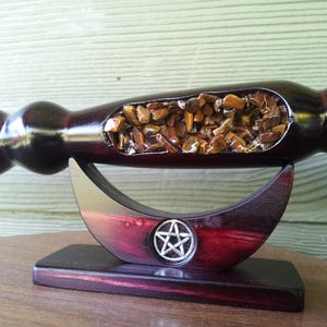 Puede incluir: Una varita de madera oscura con un acabado pulido, con una secci&oacute;n ahuecada llena de peque&ntilde;as piedras marrones y doradas. La varita descansa sobre un soporte en forma de media luna con un emblema de pentagrama.