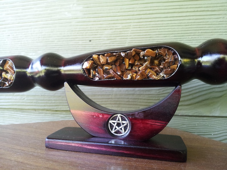 Puede incluir: Una varita de madera oscura con un acabado pulido, con una secci&oacute;n ahuecada llena de peque&ntilde;as piedras marrones y doradas. La varita descansa sobre un soporte en forma de media luna con un emblema de pentagrama.