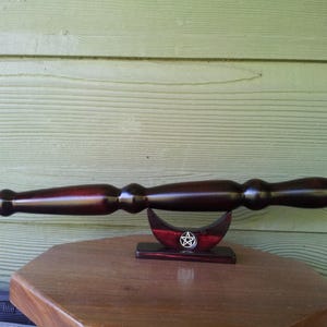 Puede incluir: Una varita de madera oscura con punta y mango bulboso descansa sobre un soporte en forma de media luna. El soporte es rojo con un pentagrama plateado. La varita se exhibe sobre una mesa de madera.