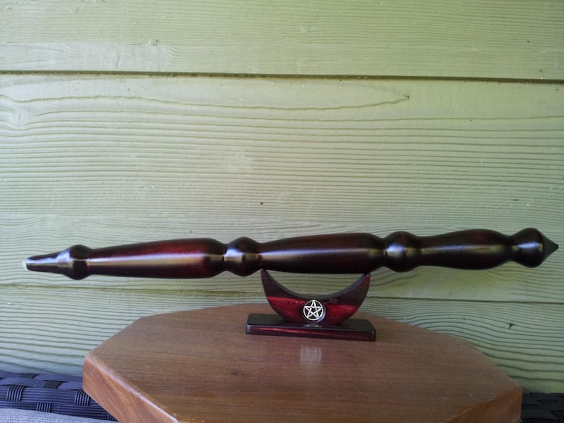 Puede incluir: Una varita de madera oscura con punta y mango bulboso descansa sobre un soporte en forma de media luna. El soporte es rojo con un pentagrama plateado. La varita se exhibe sobre una mesa de madera.